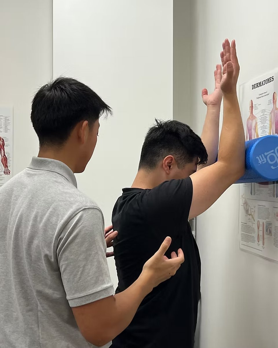 A physiotherapist at Wally's Health Clinics guiding a patient through an overhead shoulder-raise movement assessment against the wall in the Darling Square clinic.